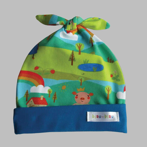 baby hat with knotted top and blue rim wtih pattern of green hills and trees, rainbows, clouds, red roofed house and smiling pig