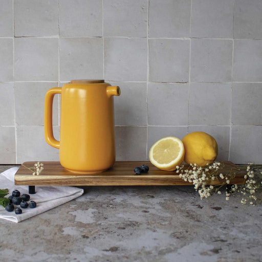 yellow teapot with long handle and short spout and cork lid. on a chopping board with cut yellow lemons and blueberries, grey tiles in the background