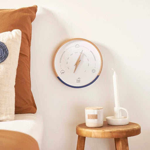 white faced wall weather barometer with wood and navy rim and wooden lightening bolt on clock face on a wall beside a bed with  tan and beige pillows