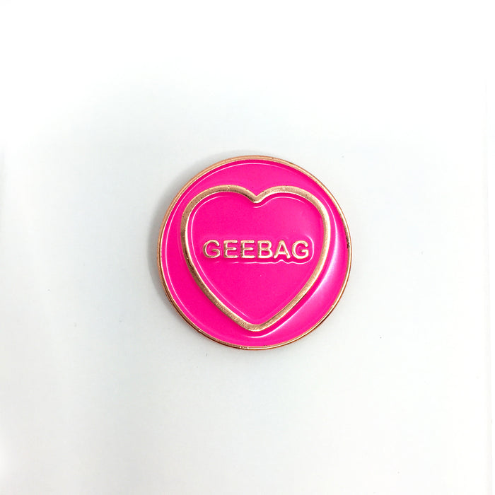 circular pink enamel pin with gold circumference, a cold heart and text in the heart that reads 'geebag' on an white background