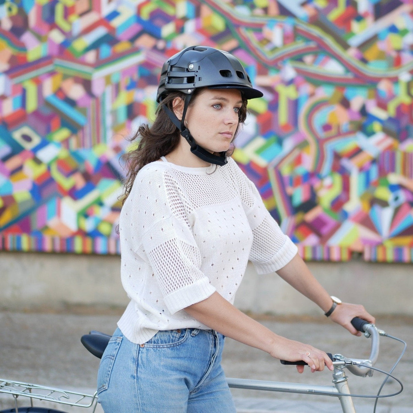 woman in white t-shirt and bicycle helmet stands against colourful wall, holding bicycle