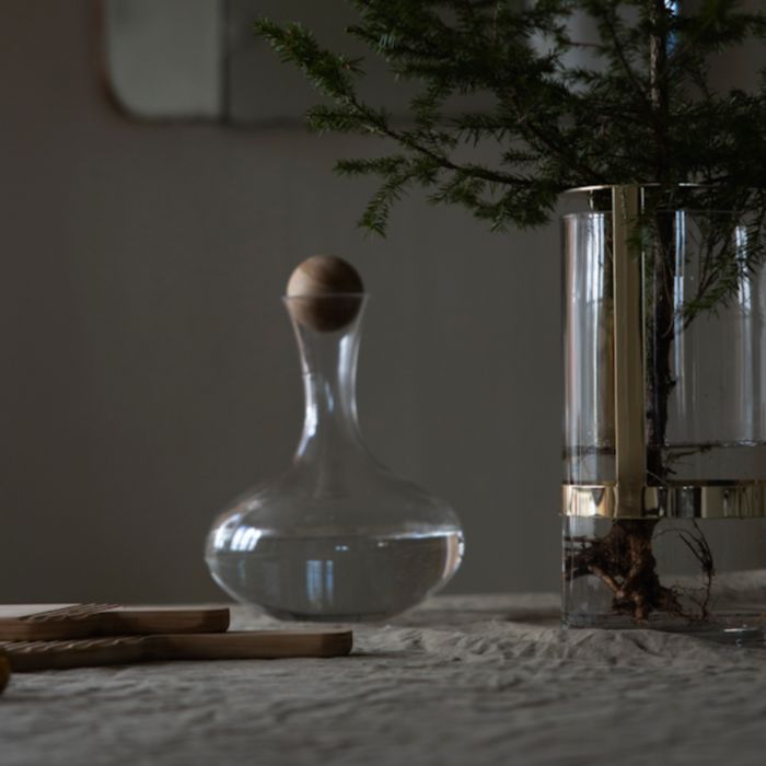 A clear glass decanter with a wooden stopper and a tall cylindrical glass vase with a gold band, holding a small evergreen tree, set on a textured tabletop in a dimly lit interior.