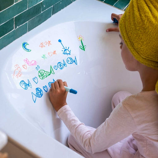 A child, in white clothes and yellow towel around head, sitting in a bath drawing pictures on the bath with a blue crayon