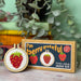 Cross stitch mini kit with completed strawberry design and packaging labeled 'I'm berry grateful' on a wooden surface with plants in the background.