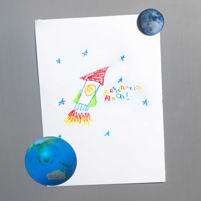 a child's crayon drawing of a rocket and stars being fixed to a metal surface by a magnet of the earth and magnet of the moon
