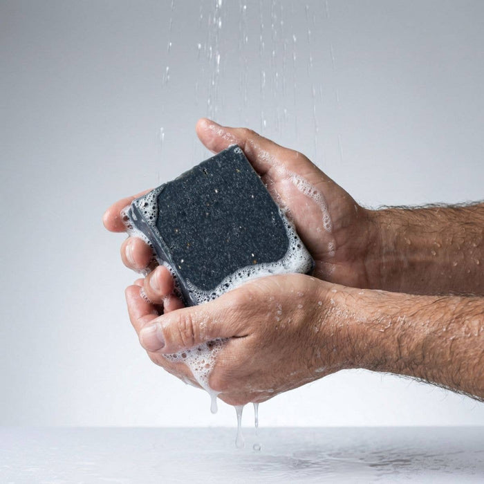 Hand holding a black soap with soap suds under running water on a light gray background