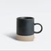 A ceramic mug with a matte black upper body and handle, and a textured beige lower band, shown on a clean light gray background.