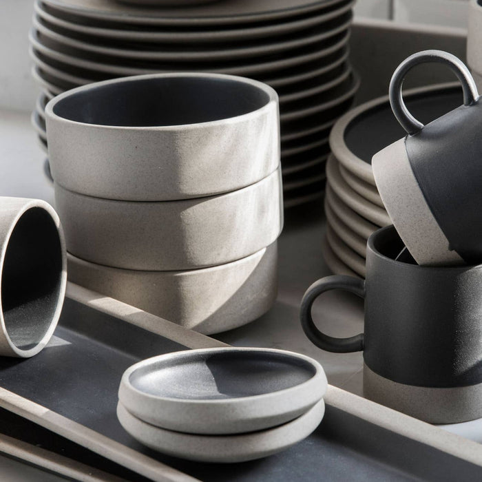 A collection of matte ceramic tableware in neutral tones, including stacked plates, bowls, mugs, and a small tray, arranged on a light surface with soft natural shadows.