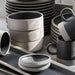 A collection of matte ceramic tableware in neutral tones, including stacked plates, bowls, mugs, and a small tray, arranged on a light surface with soft natural shadows.