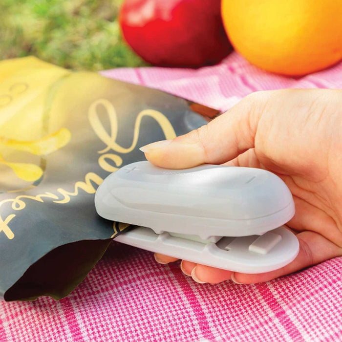 HAND HOLDING A GREY PLASTIC BAG SEALER AND CUTTER TO A BAG OF CRISPS TO SEAL THEM. THE BAG IN ON A PICNIC BLANKET WITH GRASS AND FRUIT IN THE BACKGROUND