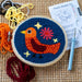 Colourful thread bird punch needle kit in a wooden hoop ring with loose thread around the exterior and a needle with blue handle grip to the right along with instruction leaflet