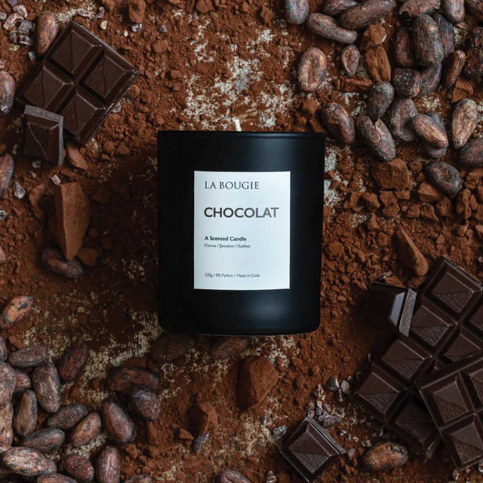 chocolat la bougie candle in a black glass container with white label laying on crushed chocolate powder, coca beans and chocolate squares