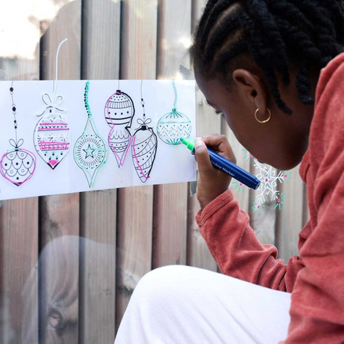 little girl with braids and gold earrings drawing on glass with a green marker. there is a template she is copying on the other side of the glass