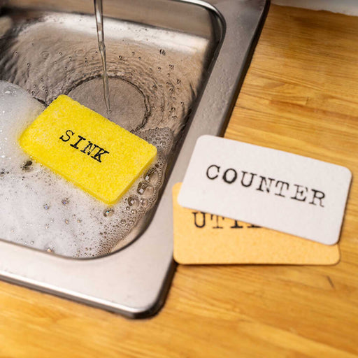 one yellow sponge in a kitchen sink and counter with running water from tap. on the counter are a white and orange sponge. all 3 sponges have text on them