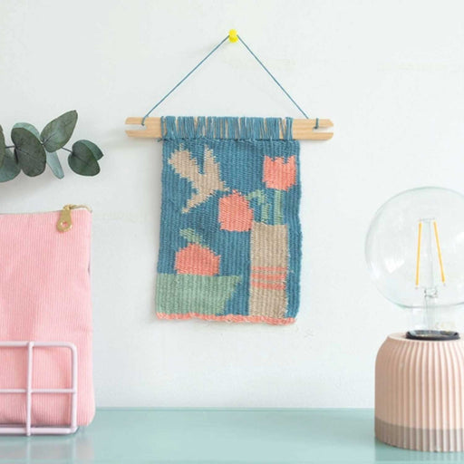 woven blue, cream and pink thread wall hanging of a flower in vase and apple in bowl, on a wall with a pink cord bag to left and lightbulb lamp to right 