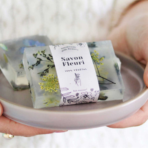holds holding a shallow grey plate with two bars of clear soap that have flowers and foiliage in the bars. The soap in front also has a white paper wrap