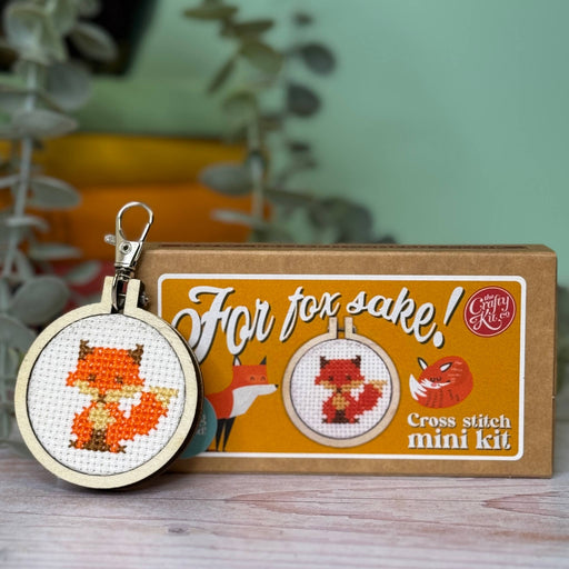 Cross stitch mini kit box with completed fox key ring on a wooden surface.