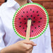 a child holding a circular pleated folding paper fan made of paper and a wooden handle, with a watermelon design on the fan