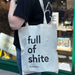 white canvas tote bag on a persons shoulder, with full of shite writen on the tote bag in black lowercase text