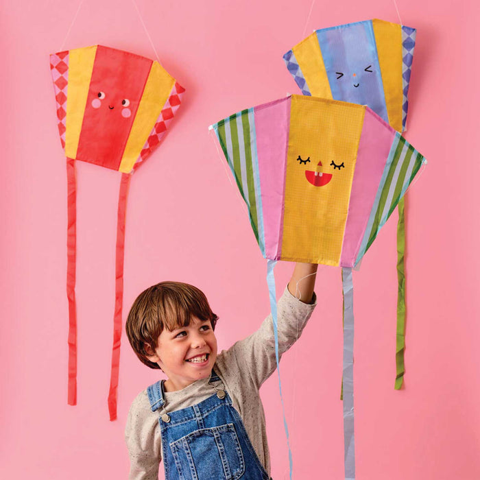 small boy with brown hair and wearing Demin dungarees smailing and holding up a fikte with a smiling face with two other kites hanging on a pink wall in the background