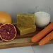 stack of 2 bars of soap with poppy seeds in them on a wood board with 2 carrots, lass of milk and cut blood orange