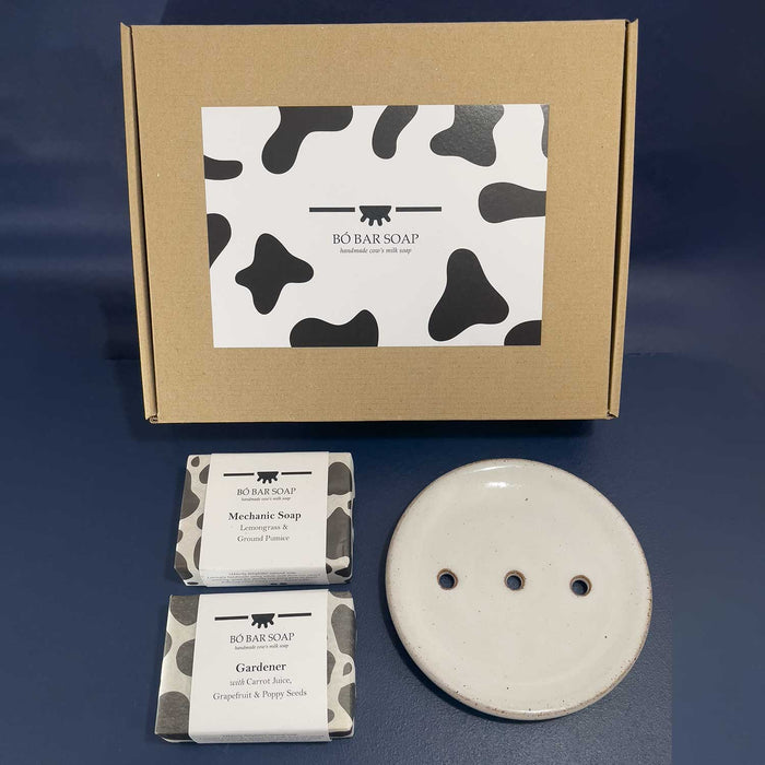 2 bars of soap and round soap dish with 3 holes in it in front of a brown  gift box with a black and white cow print label