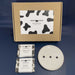 2 bars of soap and round soap dish with 3 holes in it in front of a brown  gift box with a black and white cow print label