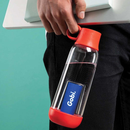hand holding a red cap and base clear water bottle. it is being held and hip height and the person is also carrying a laptop and notebook