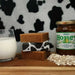 table with a lass of mink to left, 3 stacked soap bar in centre, one wrapped in cow print paper and a jar of honey with scattered oats to the right