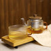 Karl Tea for One set with ridged glass cup an matching teapot with wooden lid. Both teacup and teapot have clear glass and amber glass handles, contain tea and are on a wooden serving tray