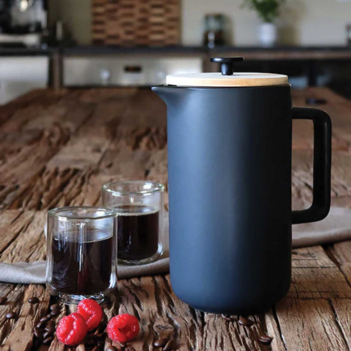 black ceramic french press with bamboo lid on a wooden kitchen table to teh right of 2 glasses of black coffee and 3 raspberries
