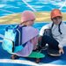 two young kids on skateboards wearing caps and blue and turquoise backpacks with yellow zips