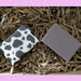 two bars of soap sitting on brown paper strips. the soap on the left is wrapped in black and white cow print paper
