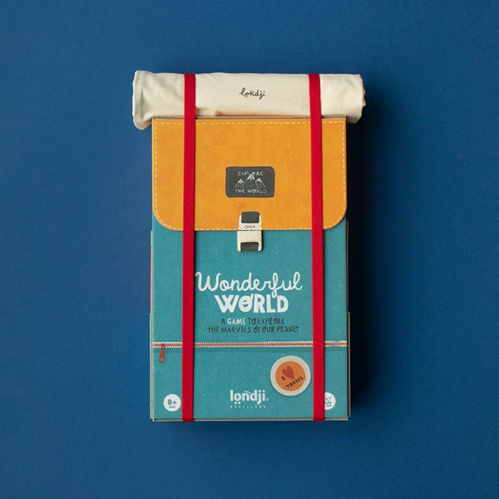 A travel-themed board game box styled like a backpack, featuring a teal lower half with 'Wonderful World' text, a mustard-yellow upper flap, red elastic straps, and a white rolled pouch on top, photographed against a solid blue background.