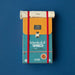 A travel-themed board game box styled like a backpack, featuring a teal lower half with 'Wonderful World' text, a mustard-yellow upper flap, red elastic straps, and a white rolled pouch on top, photographed against a solid blue background.