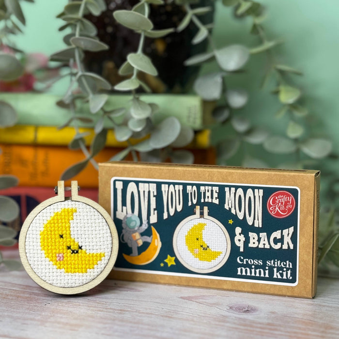 Cross stitch mini kit with 'Love You to the Moon & Back' design on a wooden surface with plants in the background.