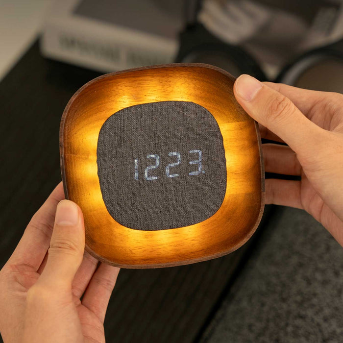 Lumos Clock, a square dark walnut wood clock with grey fabric digital clock face. The wooden frame of the clock is illuminated from behind the clock face. The clock is being held in someone's hands