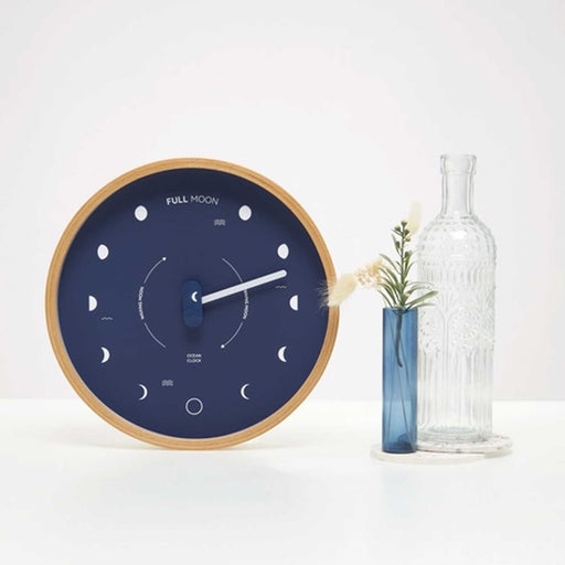 Lunar Phase Clock with blue clock face and white moon pattern to right of clock is a blue glass vase with foliage and a clear glass bottle