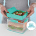 woman hands and torso in grey top holding a teal lunch box comparment filled iwth salad over the a clear plastic lunchbox with fruit and a bread roll inside it