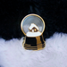 Pin with a snow globe design featuring a house on a fluffy white surface with a dark background