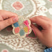 child's hand threading pink thread though a wooden flower template with thread holes