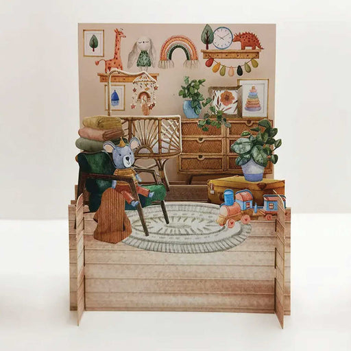 pop out greeting card showing a baby's bedroom with chair and blue teddy, two houseplants toy train, green rug and wicker cot