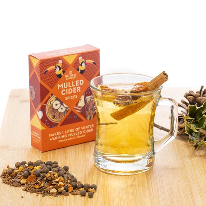 a glass cup of mulled cider with stick of cinnamon in it and orange mulled cider spice box to left with loose spices in front of it