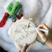 Hands holding an embroidery ring with a mushroom line drawing in canvas in it. They are using a needle tool to punch green thread in the canvas with various coloured threads on a blanket beside the embroidery ring