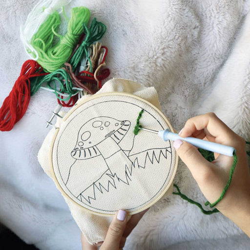 Hands holding an embroidery ring with a mushroom line drawing in canvas in it. They are using a needle tool to punch green thread in the canvas with various coloured threads on a blanket beside the embroidery ring