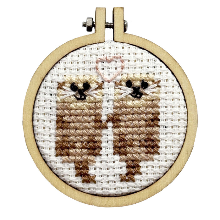 Cross-stitched design of two otters holding hands in a wooden hoop on a white background