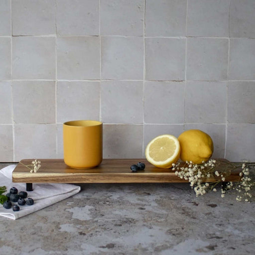 yellow cup with no handle on a wooden tray to the left of 2 lemons, white flowers and blueberries