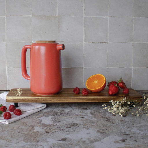 RED COLOURED TEAPOT WITH LONG HANDLE AND SHORT SPOUT AND CORK LID. ON A WOODEN BOARD WITH HALF AN ORANGE AND RASPBERRIES