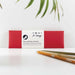 red pencil pouch with 3 pencils on table in front. Pencil case has a white cardboard wrap and there are green fronds in the foreground