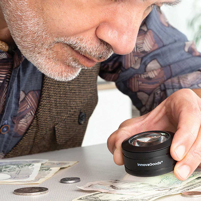 man in blue shirt and brown waistcoat with white stubble on face hold a black Pocket Magnifier with Led Light above money notes on a table
